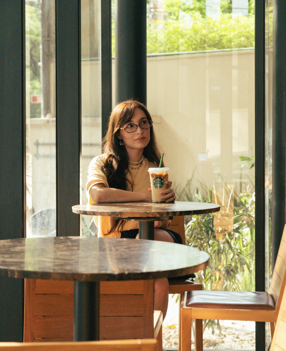Nicole Caroline sentada em um café, segurando um copo de bebida gelada perto da janela.